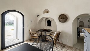 Una habitación con suelo de piedra, paredes blancas y una mesa negra con sillas. Hay dos ventanas pequeñas y una puerta que da al exterior.