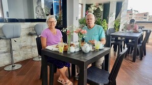 Un comedor con una mesa dispuesta para dos personas, con un jarrón de flores, una tetera y un vaso de zumo de naranja.
