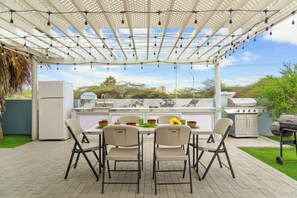 An outdoor patio with a pergola, string lights, a grill, and a table with chairs.