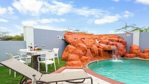 Un espace piscine agrémenté d’une cascade artificielle, de chaises longues et d’une table où sont servis des boissons.