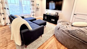A modern living room with a sofa, ottoman, and a bean bag. A flat-screen TV is mounted on the wall, and there is a door on the right. - Charming, Fully Restored 1.5 Story Farmhouse on Acreage (Muenster)