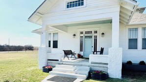 A white two-story house with a covered porch, steps leading to the entrance, and two outdoor seating areas. - Charming, Fully Restored 1.5 Story Farmhouse on Acreage (Muenster)