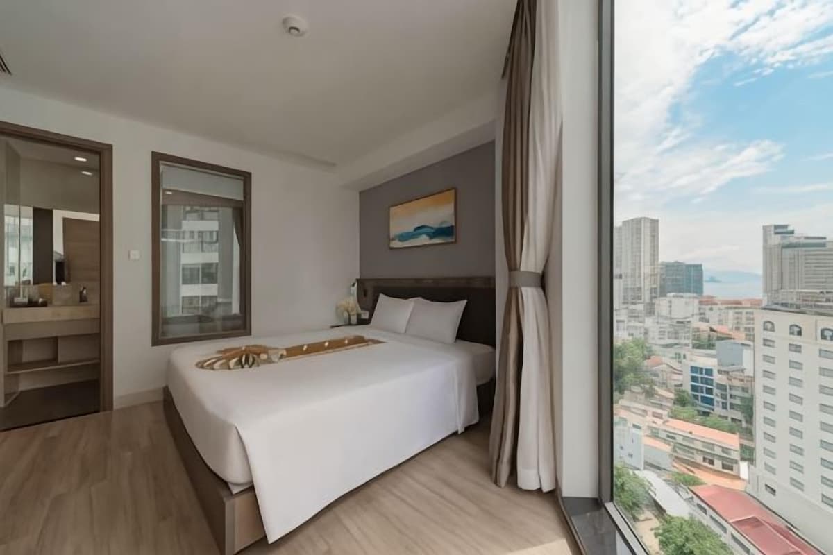 A hotel room with a bed, a large window with a city view, a painting on the wall, and a mirror.
