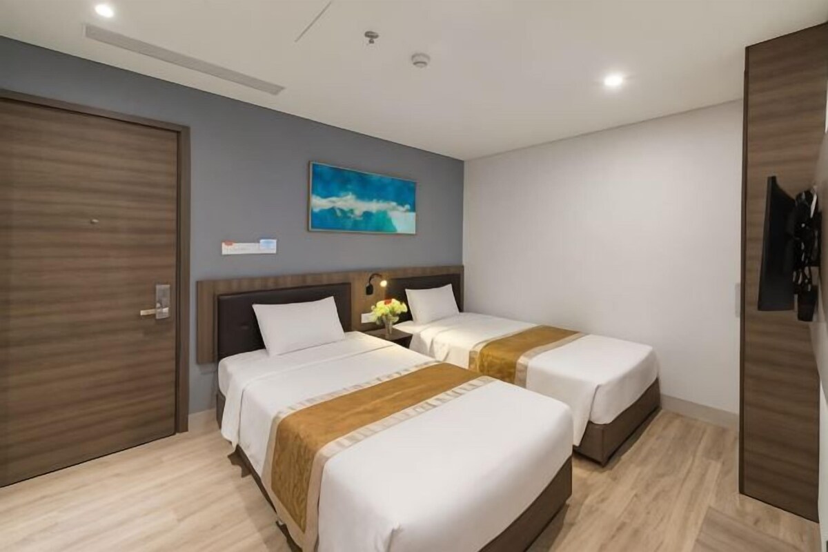 A hotel room with two beds, a wooden wardrobe, a painting on the wall, and a nightstand with a lamp.