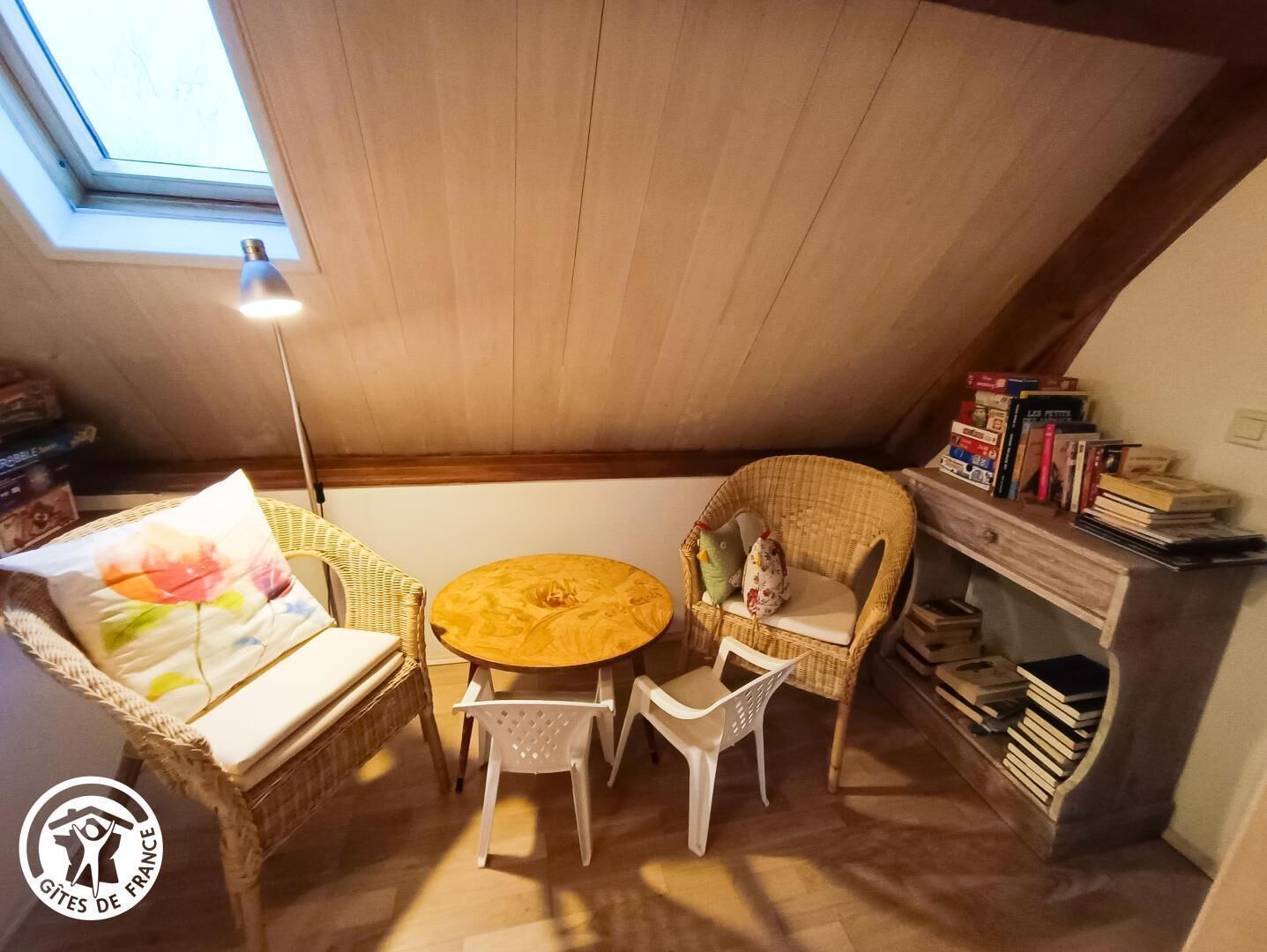 Een gezellige zolderkamer met een ronde houten tafel, rieten stoelen en een boekenplank vol boeken.
