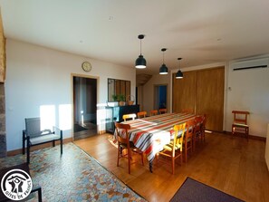 Un comedor con una mesa larga, sillas, lámparas colgantes, un reloj y una pequeña cocina con un fregadero y encimera.