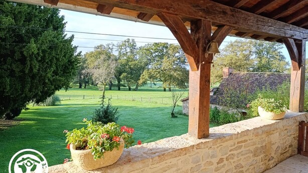 Un espacio al aire libre cubierto con paredes de piedra y vigas de madera, con plantas en macetas y vistas a un campo verde y árboles.