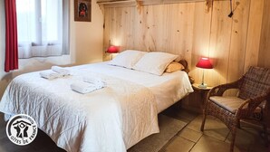 Un dormitorio con cabecera de madera, dos lámparas de noche, una cama con ropa blanca, una silla y una ventana con cortinas rojas.