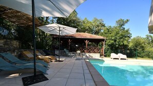 Zona de piscina con sillones de descanso, un espacio de estar cubierto y una pared de piedra.