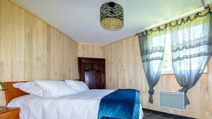 Un dormitorio con cama, paredes de madera, una puerta, una ventana con cortinas e iluminación en el techo.