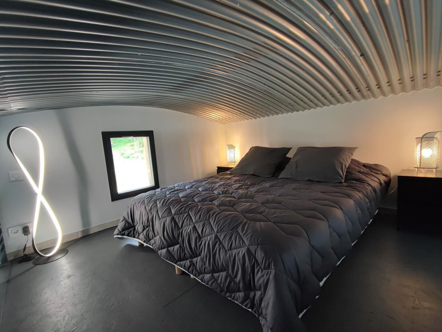Een moderne slaapkamer met een groot bed, een nachtkastje, een lamp en een raam met uitzicht op groen.