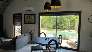 A modern living room with a sofa, dining table, and chairs, a large window with a view of a pool and greenery. - Gîtes de France® - VIP REFUGE (LE VIGNON-EN-QUERCY)