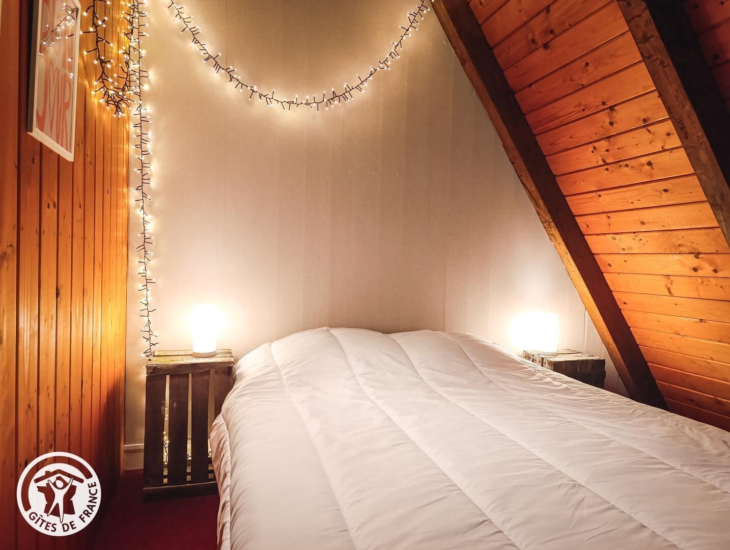 Une pièce chaleureuse avec un plafond en pente, des murs en bois, un lit avec une literie blanche et des guirlandes lumineuses.