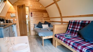 A compact, wooden interior with a kitchenette, a bed with plaid bedding, and a dining area with a white table set. - Cosy En-suite Cabin in Moray Speyside (Kinloss)