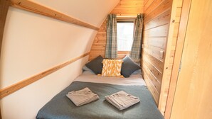 A small, cozy room with a sloped ceiling, wooden walls, a bed with a grey bedspread and two pillows, and a window with curtains. - Cosy En-suite Cabin in Moray Speyside (Kinloss)
