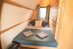Habitació petita i acollidora amb sostre inclinat, parets de fusta, un llit amb una colcha gris i dos almoinos, i una finestra amb cortines.