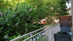 Un balcon doté d’une rambarde en métal, d’un paillasson à motifs et offrant une vue sur une végétation luxuriante et des fleurs violettes.