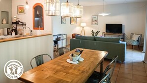 Un salon moderne avec un canapé, une table à manger et un téléviseur. Des lampes suspendues sont placées au-dessus de la table, et une lampe se trouve sur une petite table d’appoint.