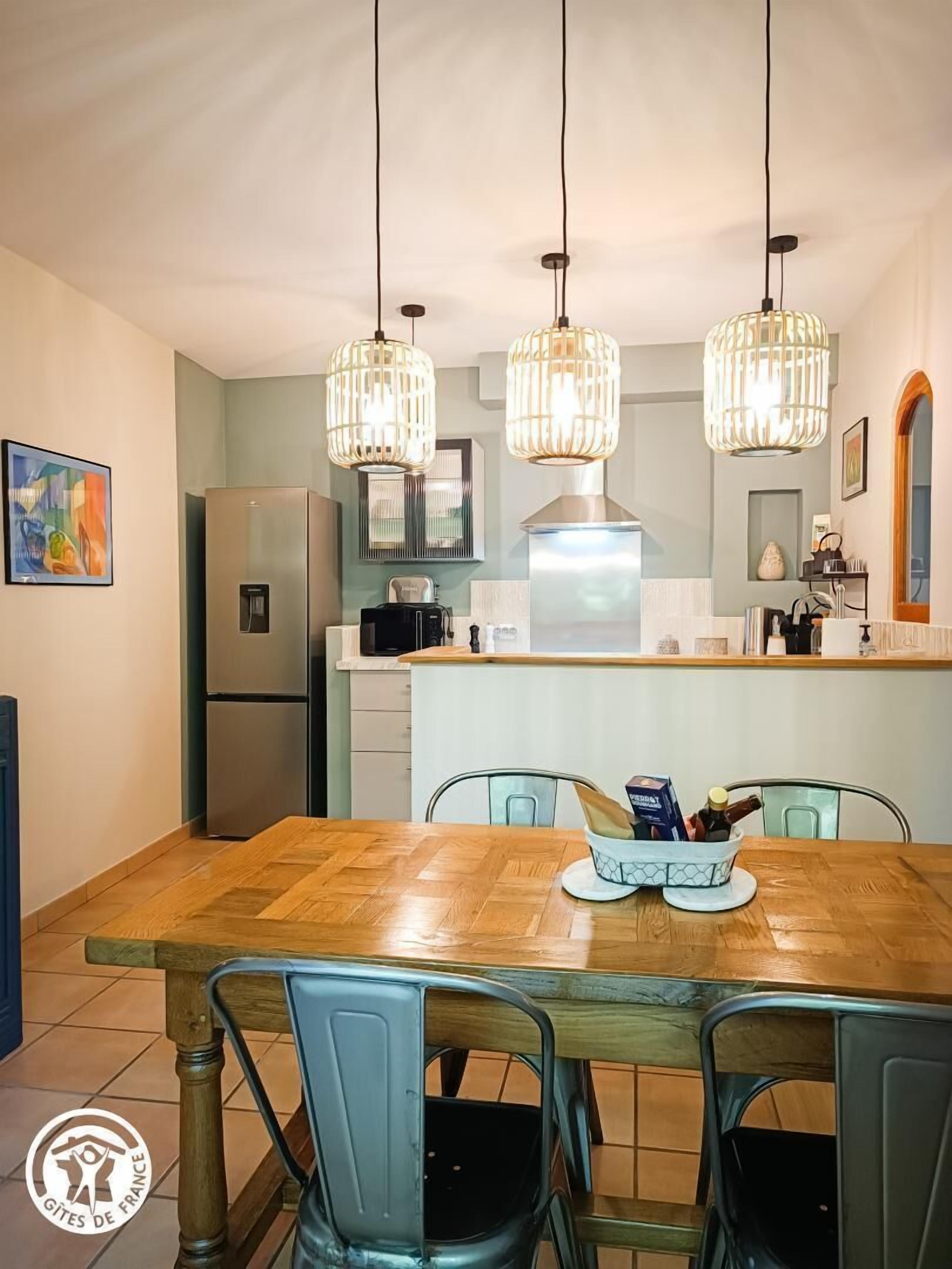 A modern kitchen with a wooden dining table, two hanging pendant lights, and a stainless steel refrigerator.