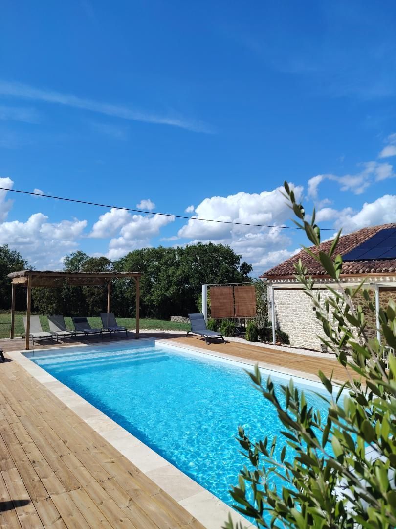 A swimming pool with lounge chairs and a pergola, surrounded by a wooden deck and a stone building.