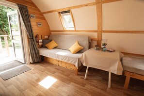 A cozy room with a wooden floor, a sofa with cushions, a small table, and a window with a view of greenery.