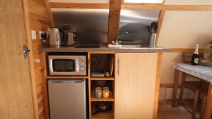 A compact kitchen with a microwave, toaster, and kettle on the countertop, a sink with utensils, and a refrigerator. - Cosy En-suite Wigwam Glamping Pod (Tenby)