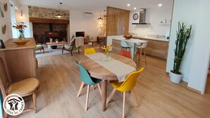 Een ruime woonkamer met een houten eettafel, kleurrijke stoelen, een keuken met een modern design en een stenen openhaard.