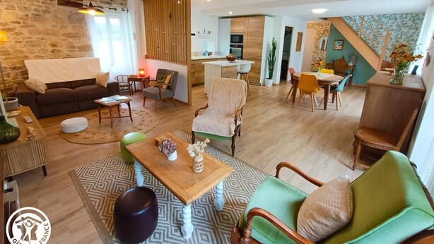 Een ruime woonkamer met een bank, salontafel en stoelen. Er is een keuken met houten kasten en een eettafel.