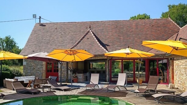 A poolside area with lounge chairs and yellow umbrellas, a stone building with a tiled roof, and a clear blue sky.