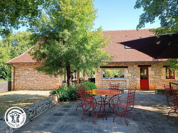 A stone building with a red door, a covered patio with a table and chairs, and a large tree in the background.