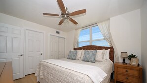 Ein Schlafzimmer mit Bett, Nachttisch, Deckenventilator und einem Fenster mit Vorhängen.