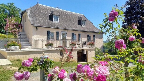 Una casa de dos pisos con balcón, un pequeño porche y un jardín con flores rosadas.