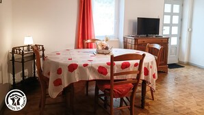 Un comedor con una mesa redonda cubierta con un mantel de motivos florales rojos, sillas de madera, un armario de madera con un televisor y una ventana con cortinas rojas.