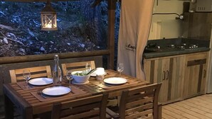 Ein Holzespeisetisch im Freien mit Tellern, Gläsern und einer Flasche, mit Blick auf Bäume und einer Küchenecke im Hintergrund.