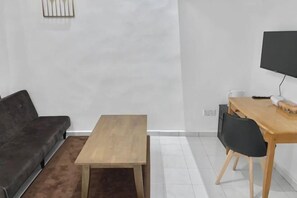 A modern living room with a dark grey sofa, a wooden coffee table, a dining table with chairs, and a flat-screen TV mounted on the wall.