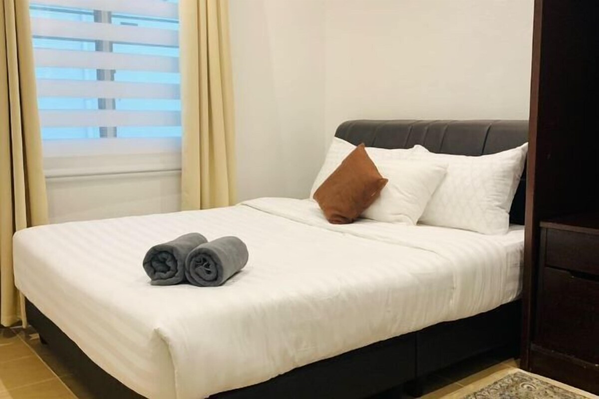 A neatly made bed with white bedding, a brown pillow, and rolled towels on the side.