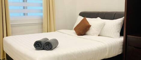 A neatly made bed with white bedding, a brown pillow, and rolled towels on the side.