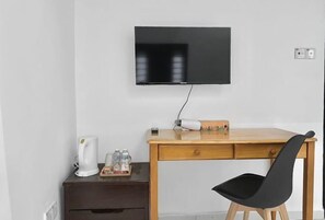 A small, tidy room with a wooden desk, a black chair, a flat-screen TV mounted on the wall, a white kettle, and a small table with a tray of items.