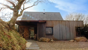 A modern building with a sloped roof, a wooden door, and a gravel pathway leading to the entrance. - A contemporary Welsh Eco house, set in an acre of land nestled on the Preseli Hills above the sea (Newport)