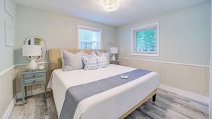 A bedroom with a bed, two bedside tables, a nightstand, a mirror, and two windows with blinds. - Scenic Drive Resort - Cabin #1 (Muskegon)