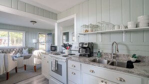 A kitchen with white cabinets, a stove, a microwave, a coffee maker, and a sink. There is a window with blinds and a door leading outside. - Scenic Drive Resort - Cabin #7 (Muskegon)
