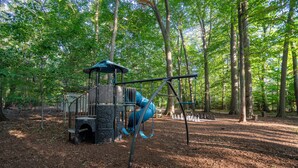 A playground with a climbing structure and swings in a wooded park. - Scenic Drive Resort - Cabin #7 (Muskegon)