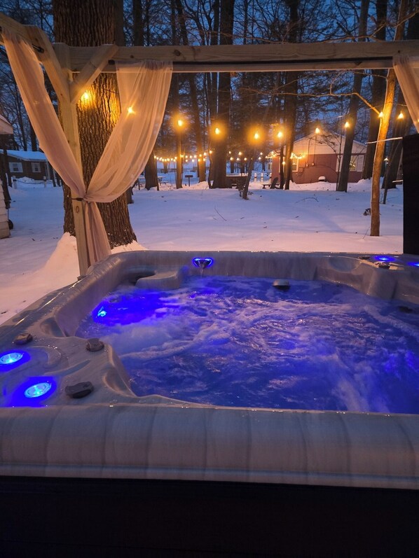 A hot tub with blue lights, surrounded by trees and snow, with string lights overhead.