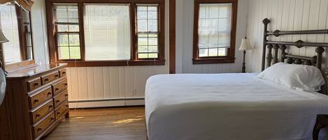 A bedroom with a large bed, wooden dresser, and two windows with blinds.