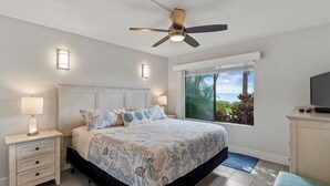 Une chambre à coucher comprenant un lit, une table de chevet, une lampe, un ventilateur de plafond, une fenêtre et un téléviseur.