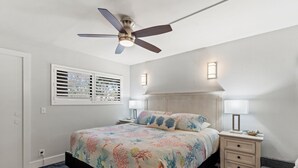 A bedroom with a bed, a ceiling fan, two bedside lamps, a nightstand, and a window with blinds.