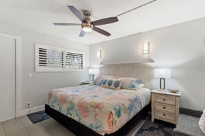 A bedroom with a bed, a ceiling fan, two bedside lamps, a nightstand, and a window with blinds.