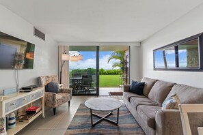 Ein modernes Wohnzimmer mit Sofa, Sessel, Couchtisch und Fernseher. Eine Glastür öffnet sich zu einer Terrasse mit Rasen und Palmen.
