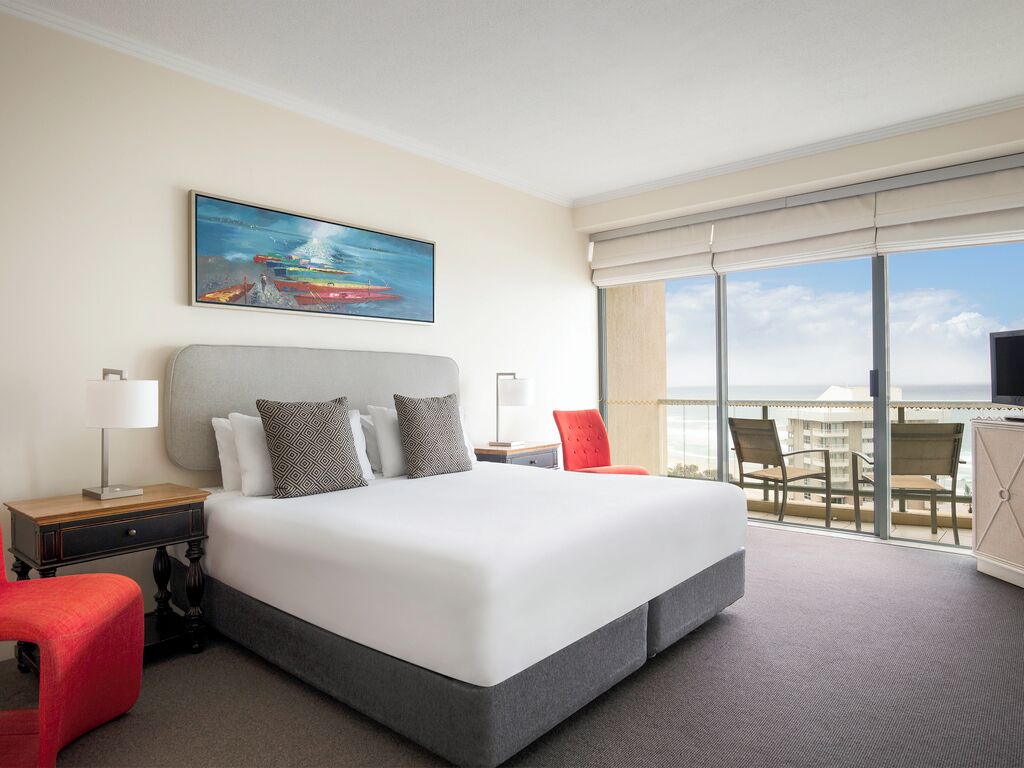 Habitación de hotel con una cama grande, un cuadro en la pared, un balcón con sillas y vista a la ciudad.
