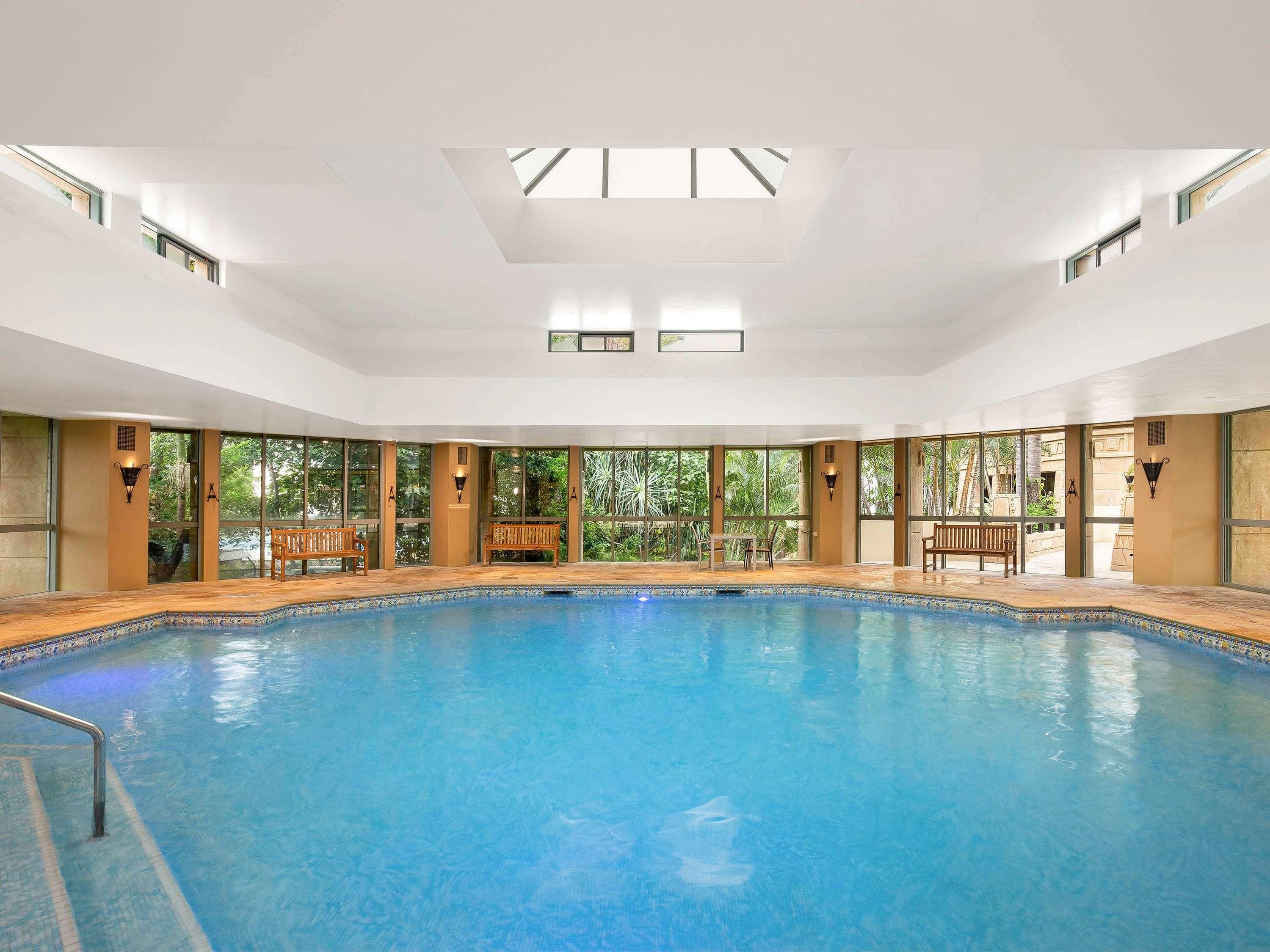 Una amplia piscina interior con paredes de vidrio transparente, bancos y un techo con tragaluces.